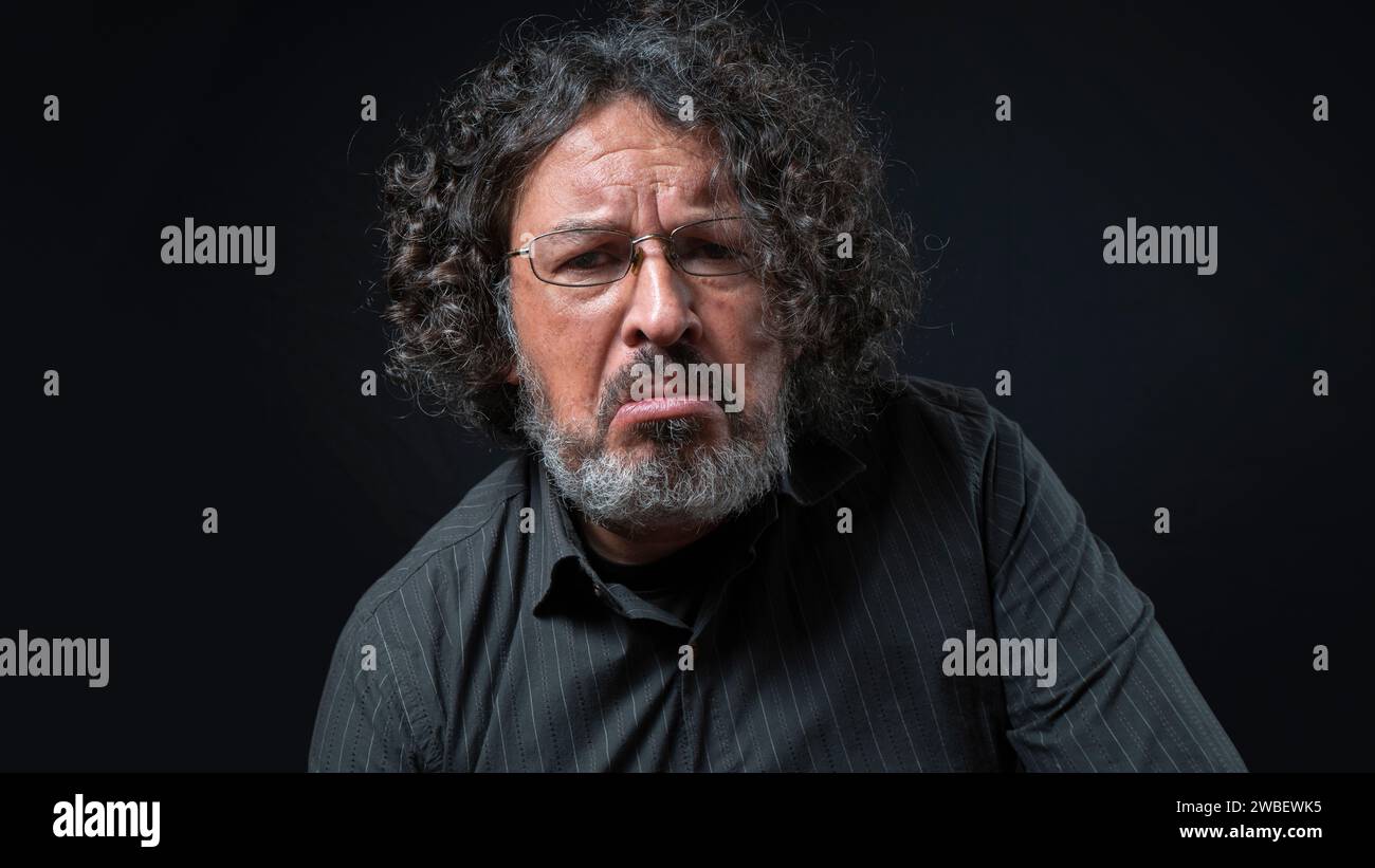 Ritratto di uomo latino con barba bianca e capelli ricci neri con triste espressione, indossando una camicia nera su sfondo nero Foto Stock Ritratto di uomo latino con barba bianca e capelli ricci neri con triste espressione, indossando una camicia nera su sfondo nero Foto Stock