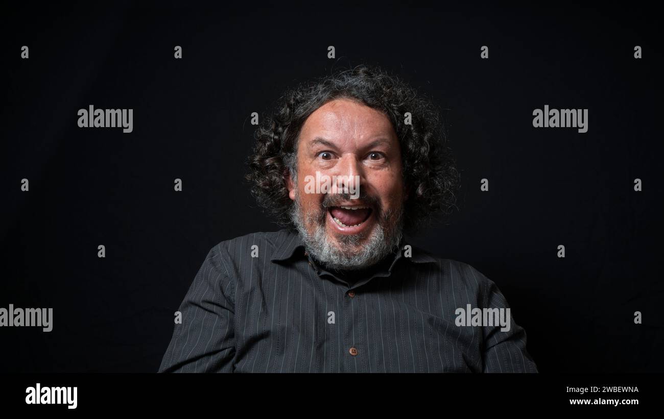 Ritratto di uomo latino con barba bianca e capelli ricci neri con un'espressione felice ed eccitata, indossando una camicia nera su sfondo nero Foto Stock