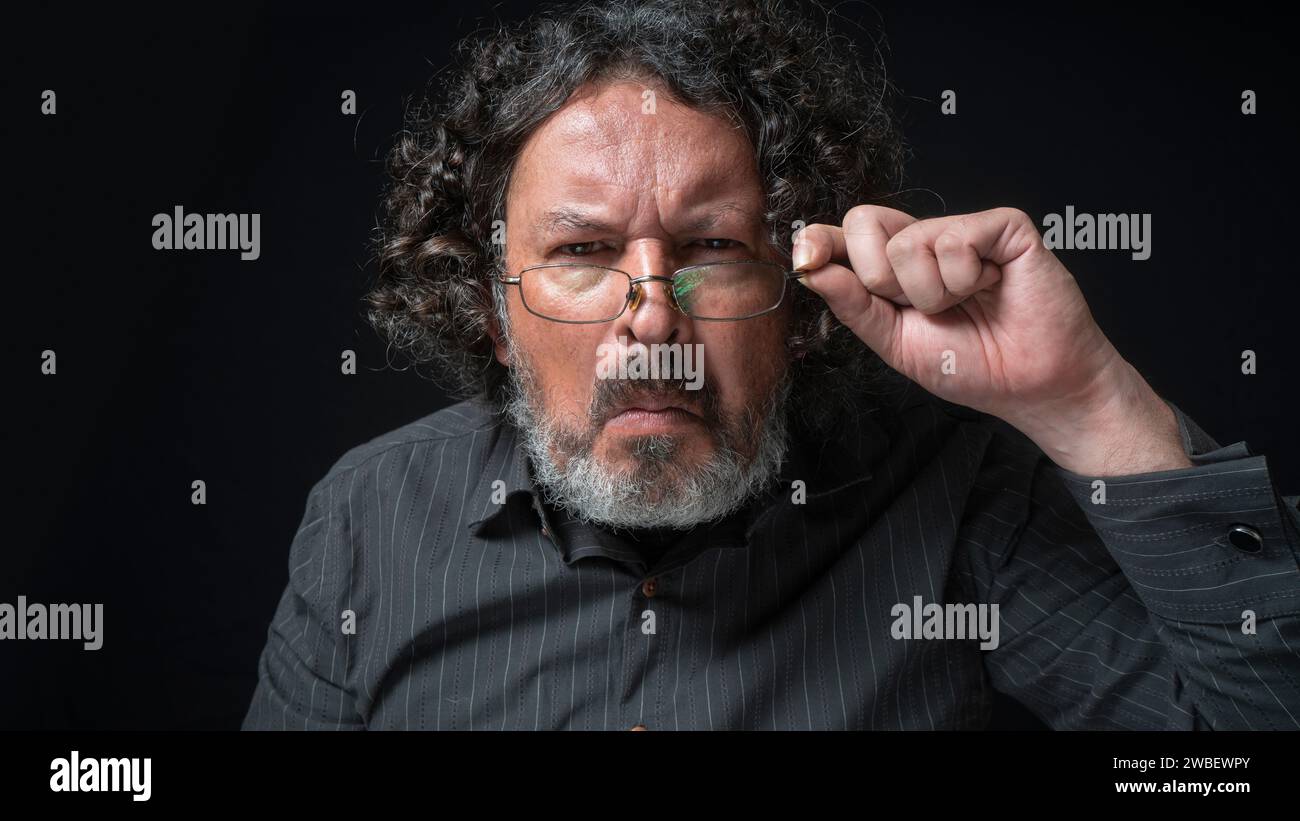 Ritratto di uomo latino con barba bianca e capelli ricci neri con curiosa espressione, guardando la fotocamera in modo sospetto, indossando una camicia nera contro il nero Foto Stock
