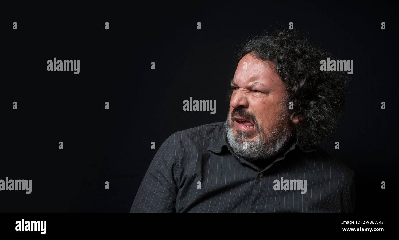 Ritratto di uomo latino con barba bianca e capelli ricci neri con espressione di rifiuto e dispiacere, guardando a sinistra, indossando la camicia nera aga Foto Stock