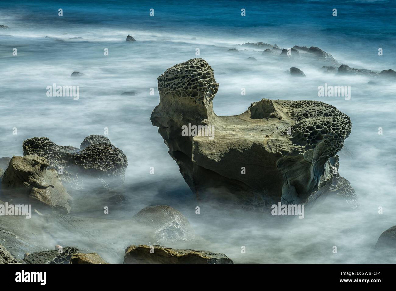 Uno scatto a lunga esposizione cattura il movimento sereno delle onde nebbiose che girano intorno alle rocce scolpite a nido d'ape in riva al mare Foto Stock