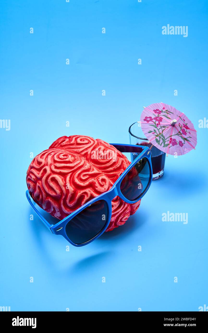 Vista dall'alto di un cervello umano di plastica rossa che indossa occhiali da sole blu accanto a un cocktail party con un ombrello su sfondo blu. Foto Stock