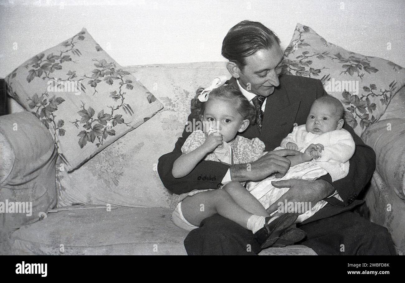 Anni '1960, un padre orgoglioso, in costume e cravatta, seduto su un divano con i suoi due figli, la figlia neonata e neonato, Inghilterra, Regno Unito. Foto Stock