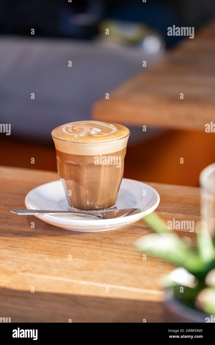 Un cappuccino appena fatto, servito in un bicchiere su un piattino in una caffetteria. Il cappuccino ha una condimento completa di crema di latte decorata con latte art Foto Stock