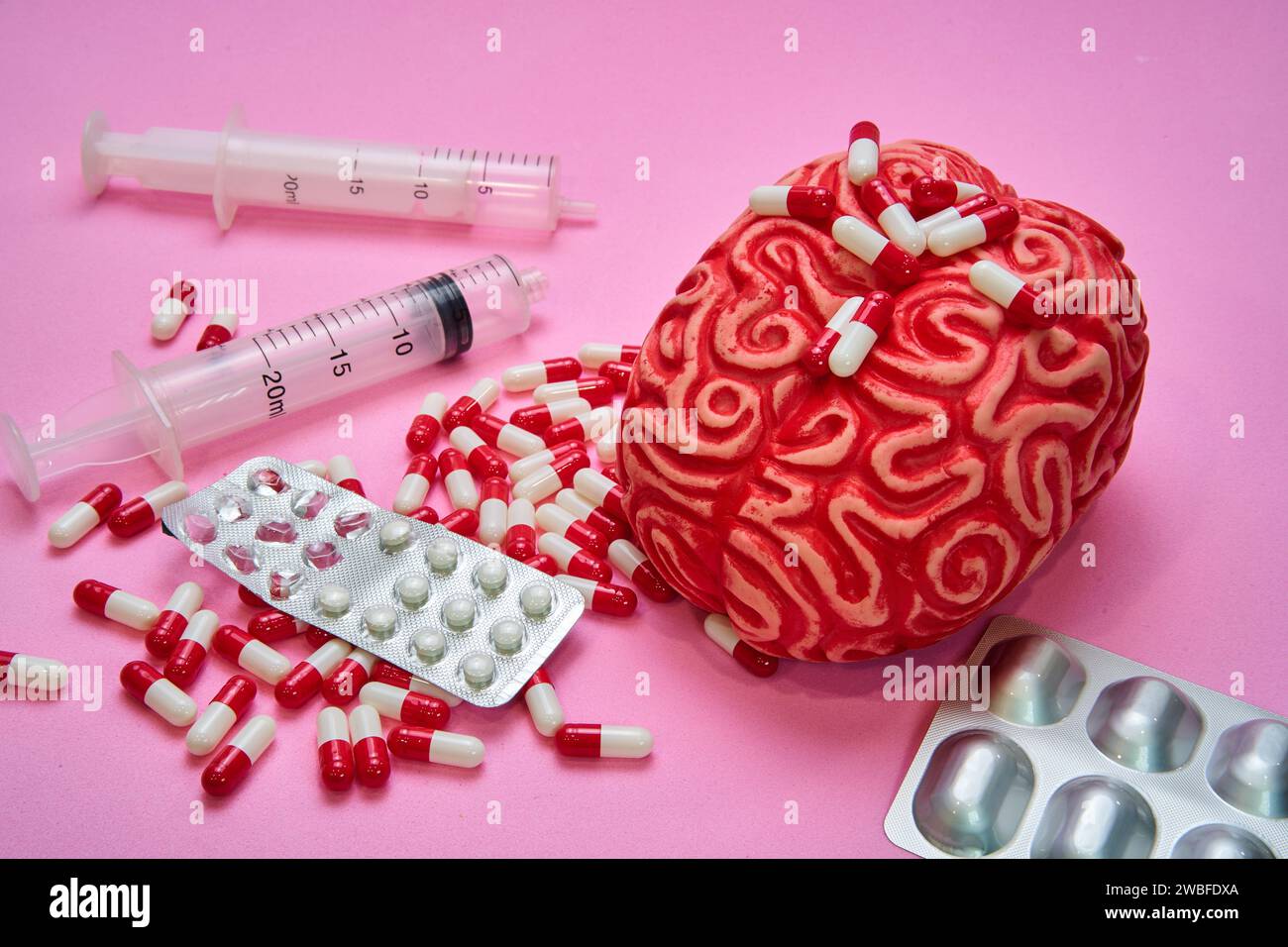 Cervello umano rosso con due siringhe e una pila di pillole e capsule rosse e bianche su un tavolo rosa. Foto Stock