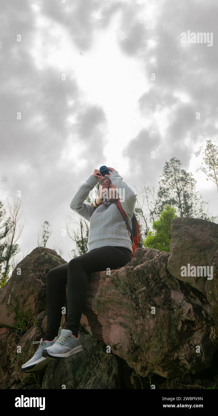 Bella giovane donna latinoamericana seduta su pietre in cima a una montagna che scatta una foto con la sua piccola macchina fotografica in una giornata nuvolosa Foto Stock