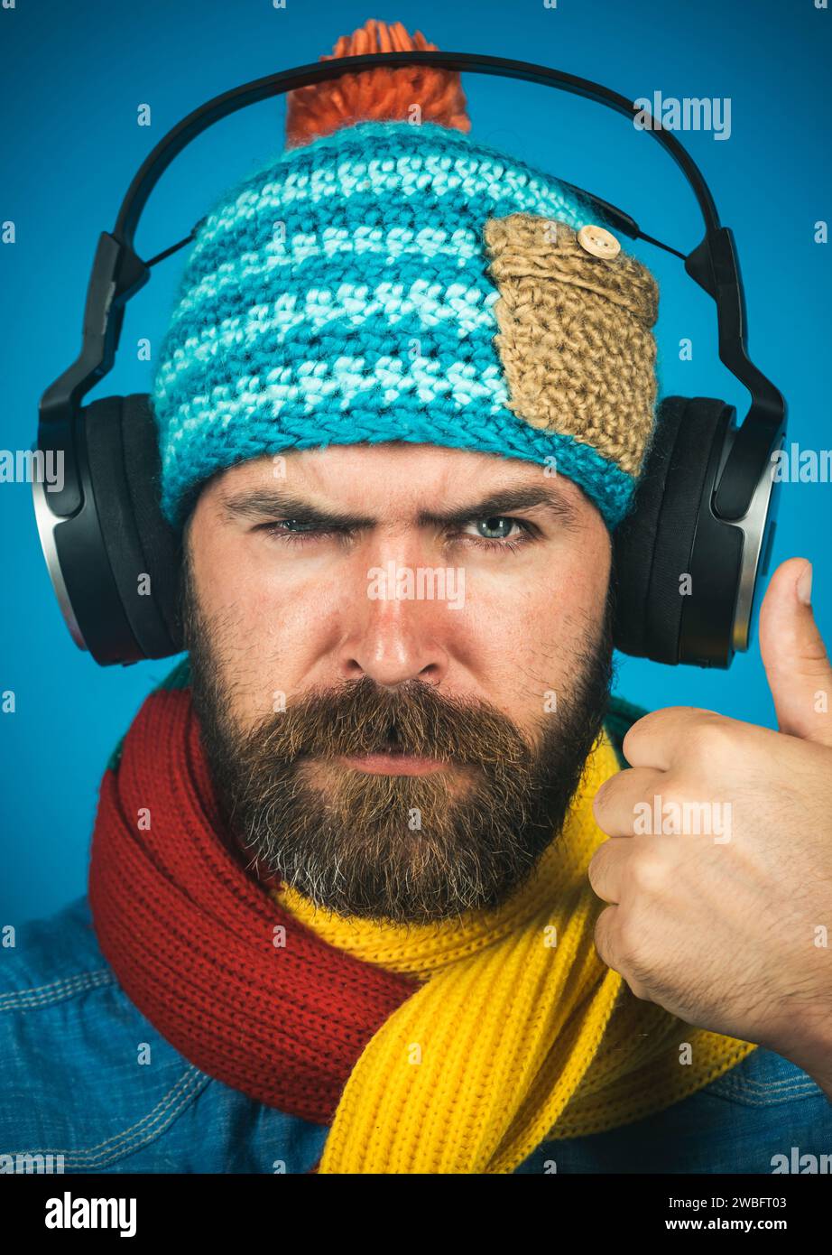 Ritratto in primo piano di un uomo barbuto serio con cappello a maglia e sciarpa in cuffie con il pollice in alto. Elegante ragazzo in camicia di denim che ascolta musica con Foto Stock