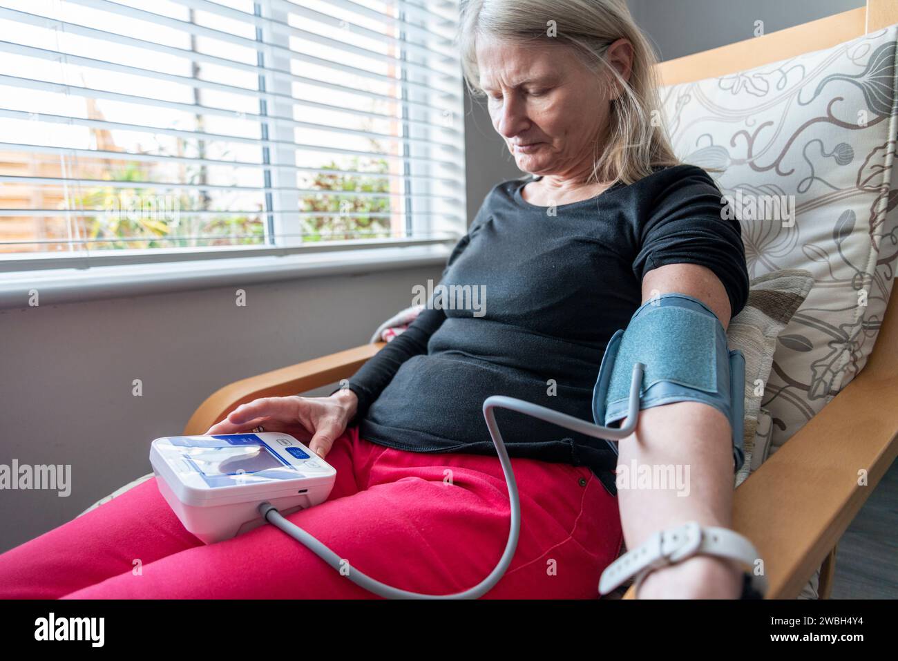 Una donna attenta alla salute, siede su una poltrona, in casa sua, usando un misuratore della pressione sanguigna per autocontrollare le sue condizioni fisiche generali, parte della sua guarigione Foto Stock