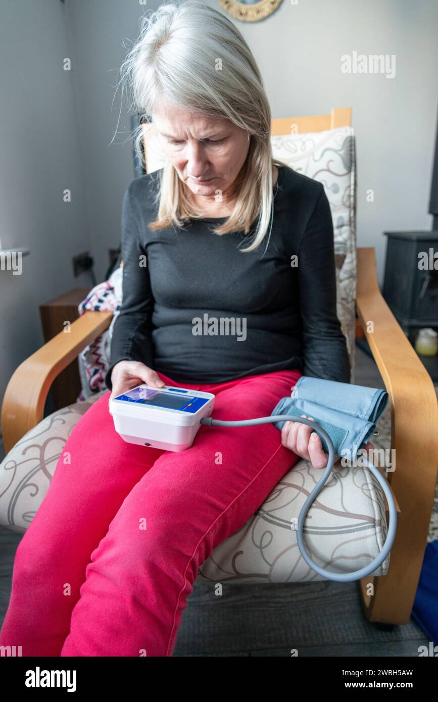 Una donna attenta alla salute, siede su una poltrona, in casa sua, usando un misuratore della pressione sanguigna per autocontrollare le sue condizioni fisiche generali, parte della sua guarigione Foto Stock