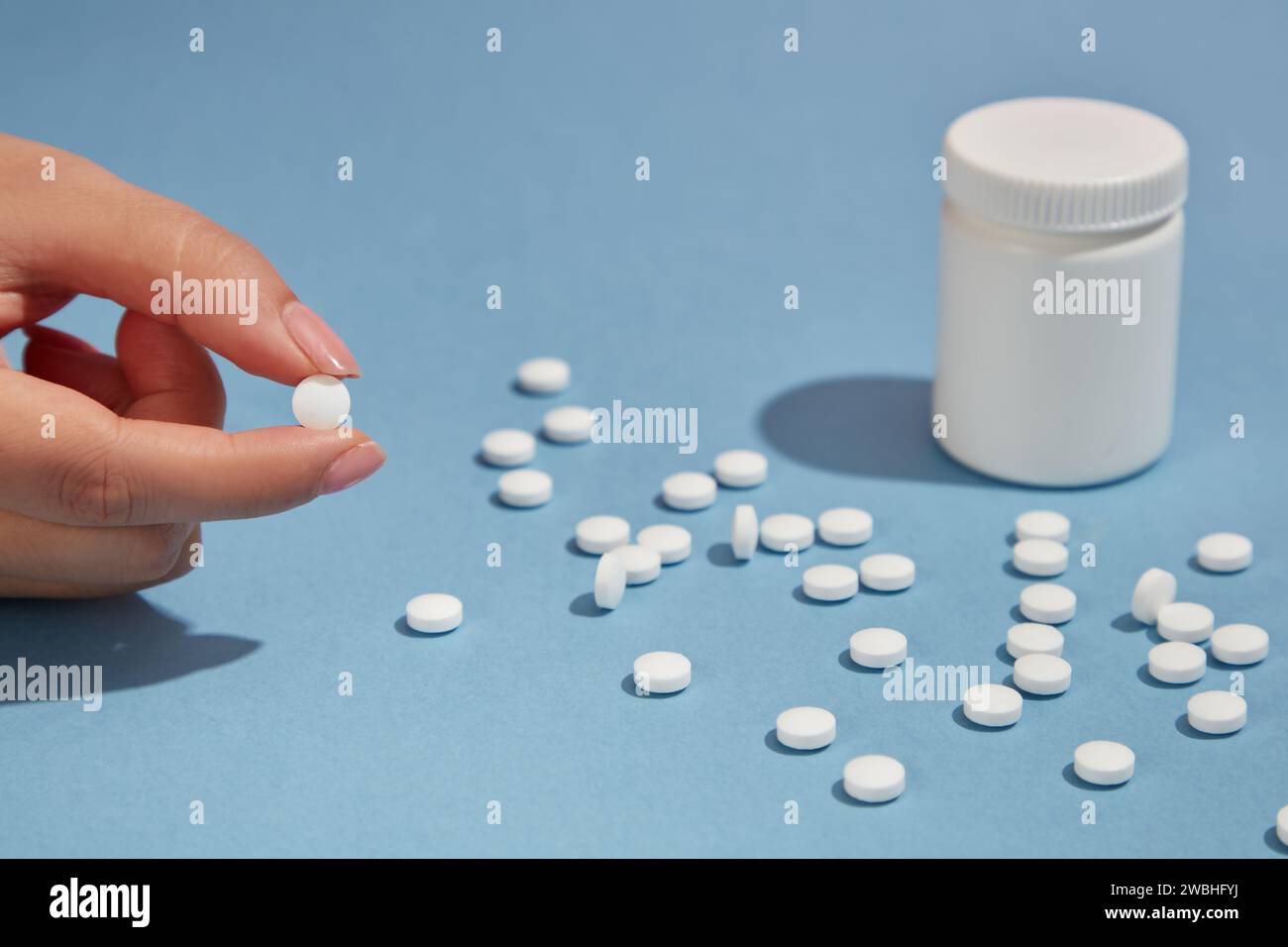 Le dita raggiungono una pillola, diverse altre pillole rotonde bianche con flacone bianco senza marchio su sfondo blu. Scena minima per la pubblicità. Selettivo f Foto Stock