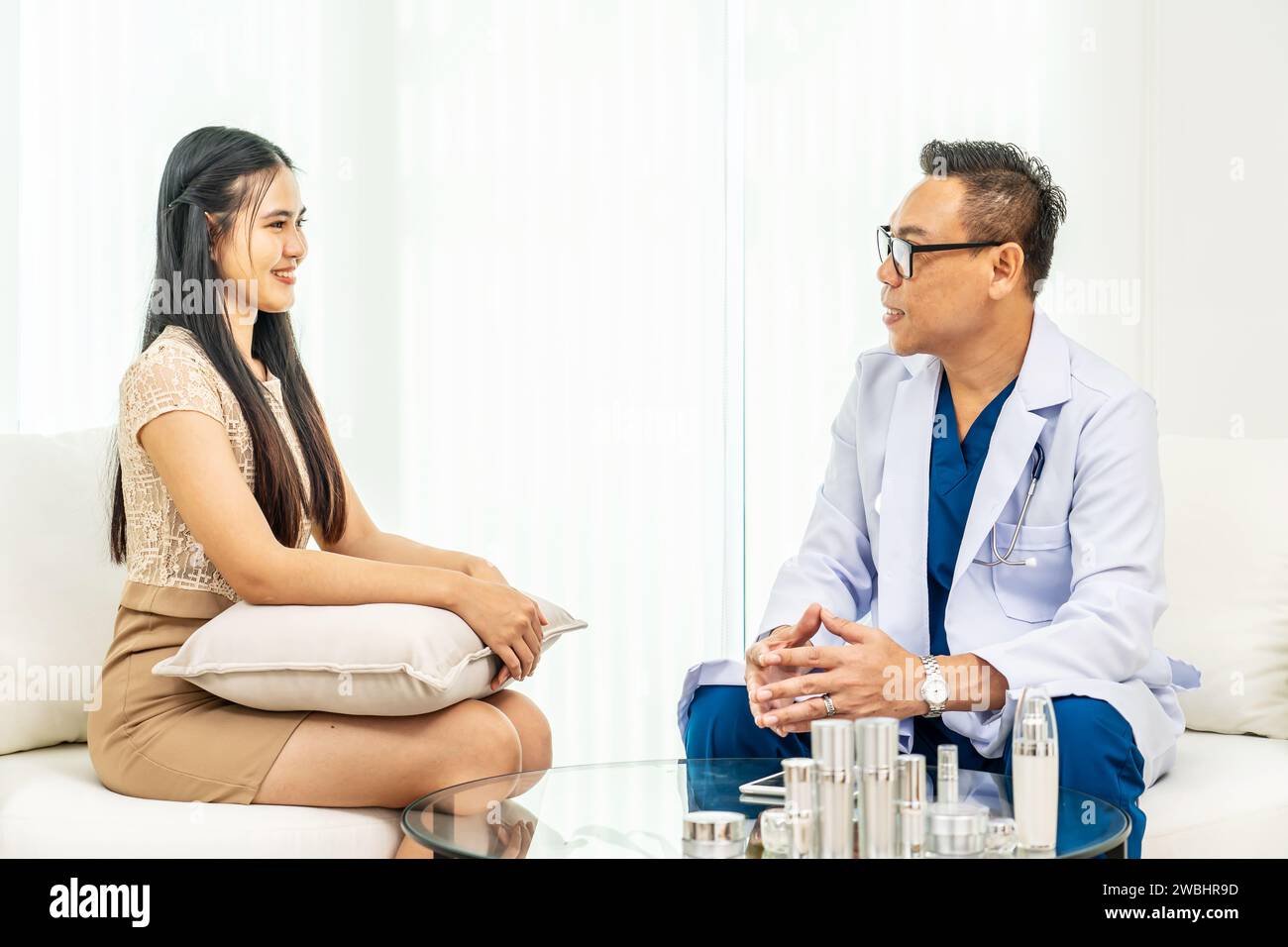 Consulente uomo con indosso abito medico che mostra creme e lozioni dalla scrivania alla donna asiatica. Dermatologo che fornisce consigli su un corretto prodotto per la cura della pelle Foto Stock