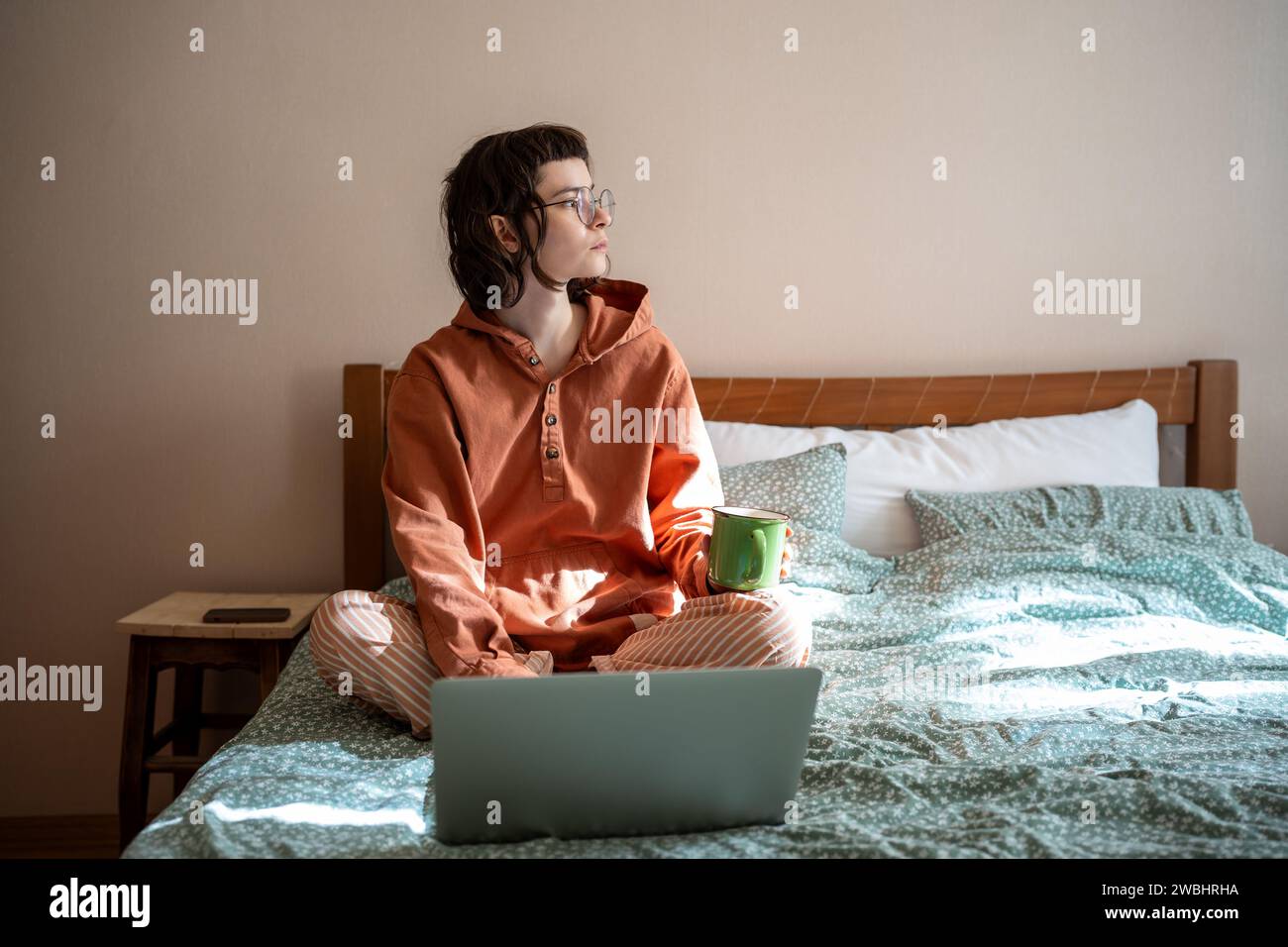 Studentessa premurosa seduta sul letto, guardando la finestra durante la pausa tè dopo aver studiato online Foto Stock