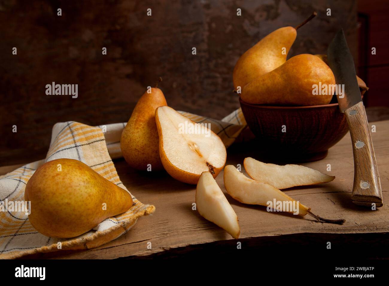 Metà e fette di frutta matura e succosa di pera e ciotola di argilla con frutta su fondo di legno vintage con asciugamano da cucina giallo. Foto Stock