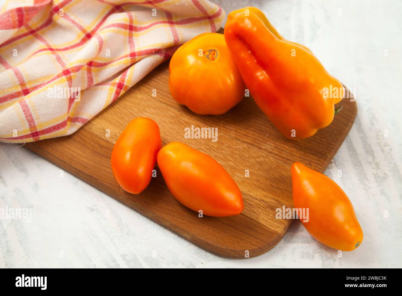 Tagliere in legno con diversi pomodori gialli succosi interi e peperoni su fondo di legno bianco con asciugamano da cucina rosso. Verdure colle Foto Stock