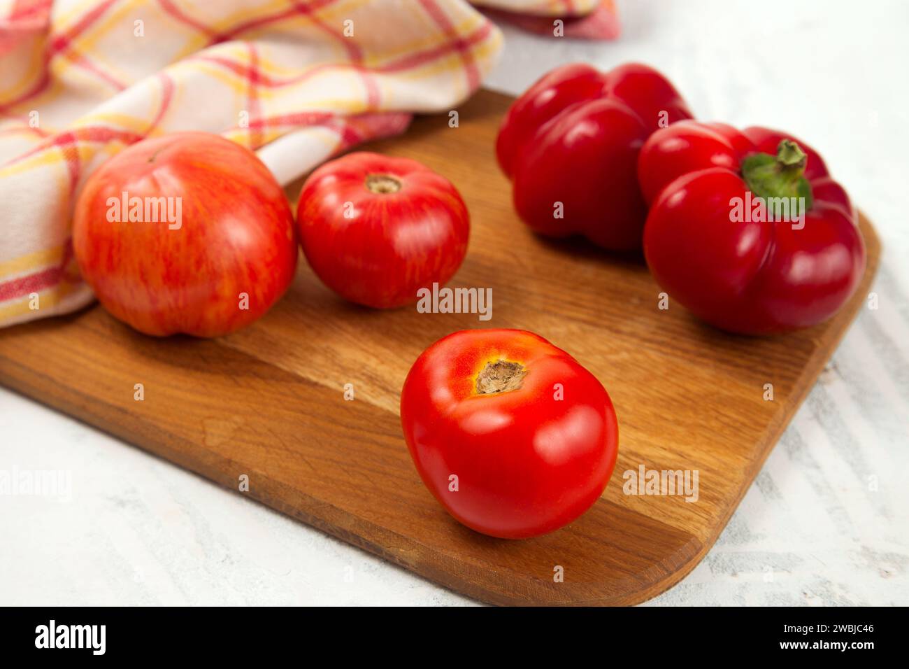 Tagliere in legno con diversi pomodori rossi succosi interi e peperoni su fondo di legno bianco con asciugamano da cucina rosso. Raccolte di verdure Foto Stock