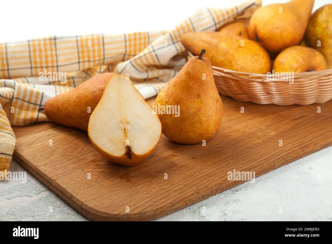 Tagliere in legno con interi e metà di succosa frutta di pera matura, cesto di vimini con pere e asciugamano da cucina giallo su fondo di legno bianco. Foto Stock