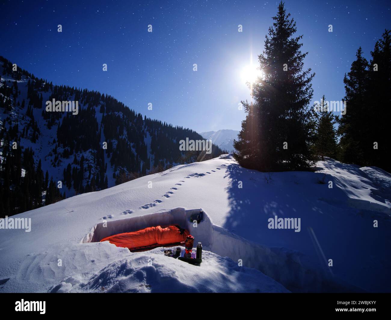 Un turista maschio dorme in un sacco a pelo in un rifugio innevato durante una notte invernale illuminata dalla luna. Escursioni di più giorni nelle montagne invernali. Sopravvivenza invernale nel Foto Stock