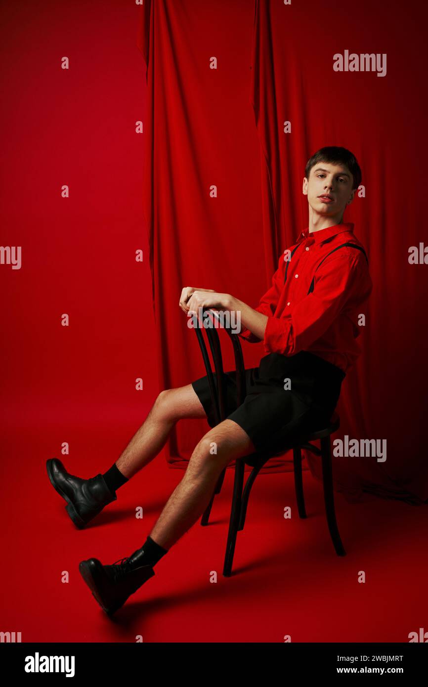 giovane uomo con camicia e pantaloncini neri seduto sulla sedia con posa sicura accanto alla tenda rossa Foto Stock