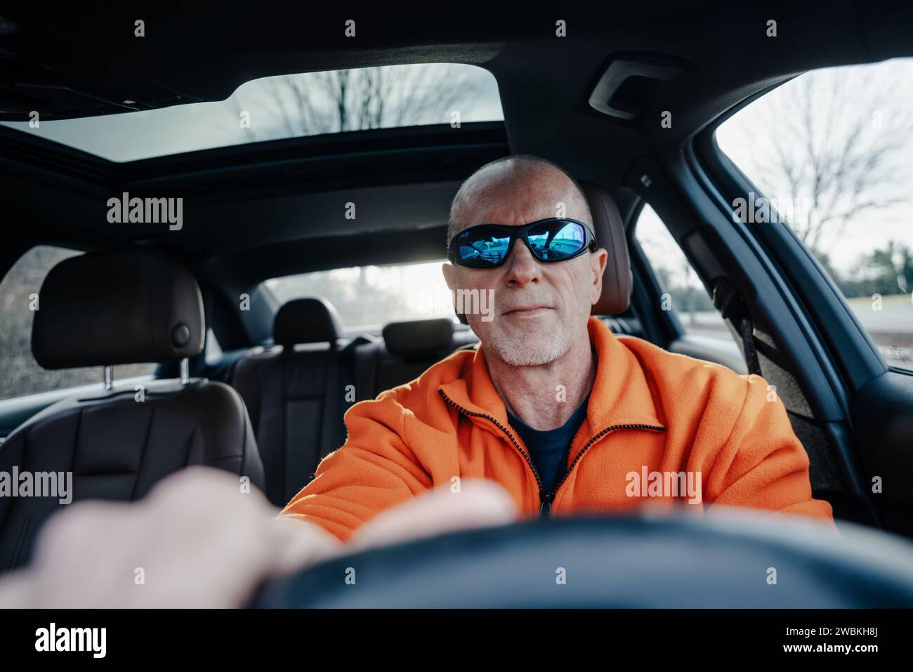 Primo piano di un uomo più anziano con occhiali da sole e giacca arancione che guida una vista interna dell'auto Foto Stock