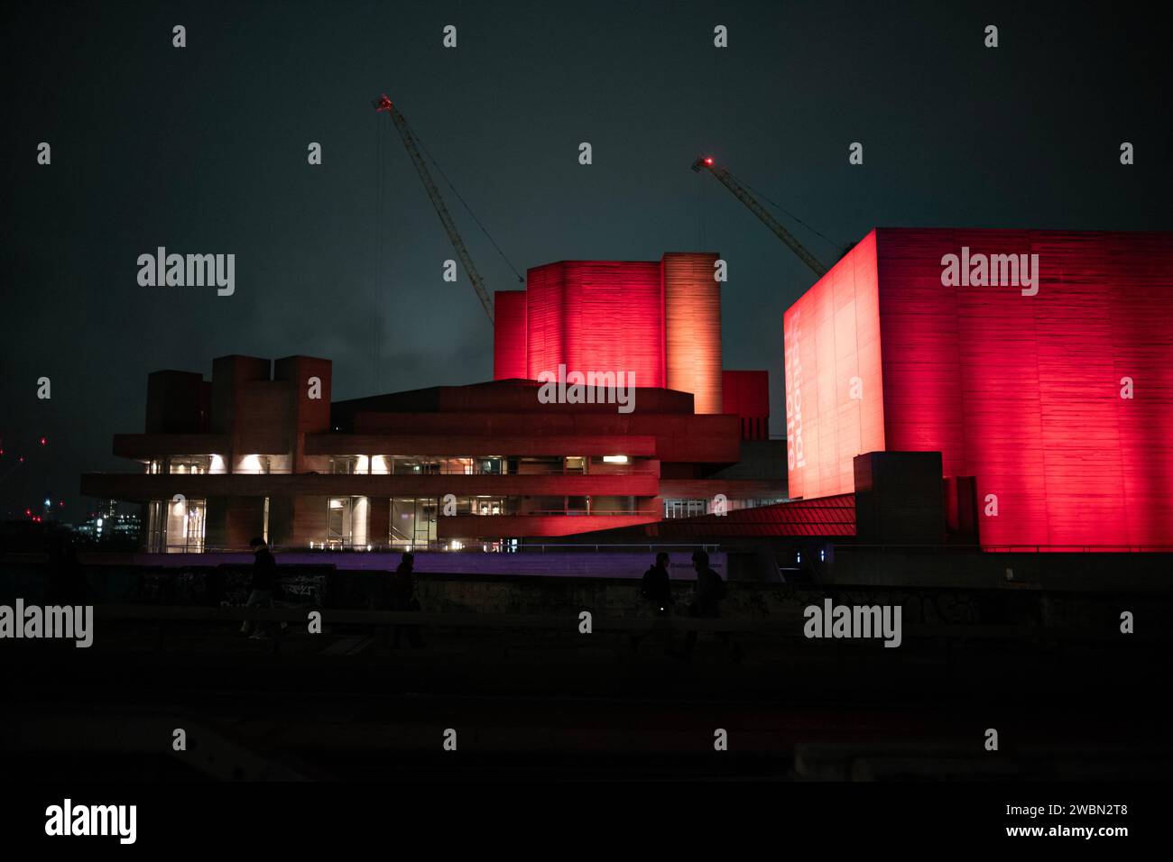 I pendolari tornano a casa in una notte d'inverno attraverso il Waterloo Bridge accanto a un National Theatre illuminato di colore rosso bankside, Southbank, nel centro di Londra, in Inghilterra. Foto Stock