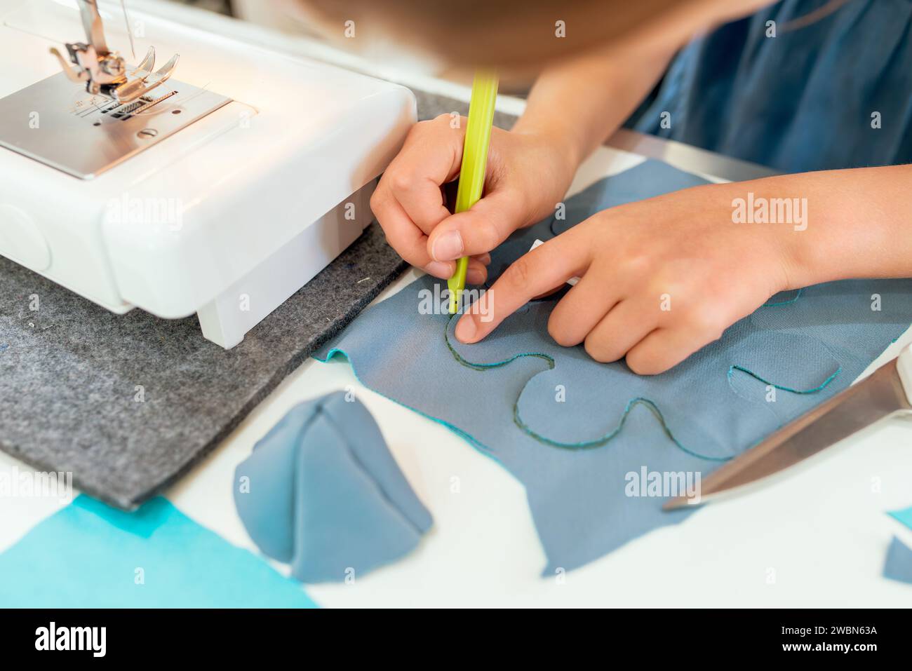 Primo piano di mani femminili che lavorano su una macchina da cucire. È seduta al tavolo e disegna con una matita verde. Laboratori creativi di sartoria per bambini Foto Stock