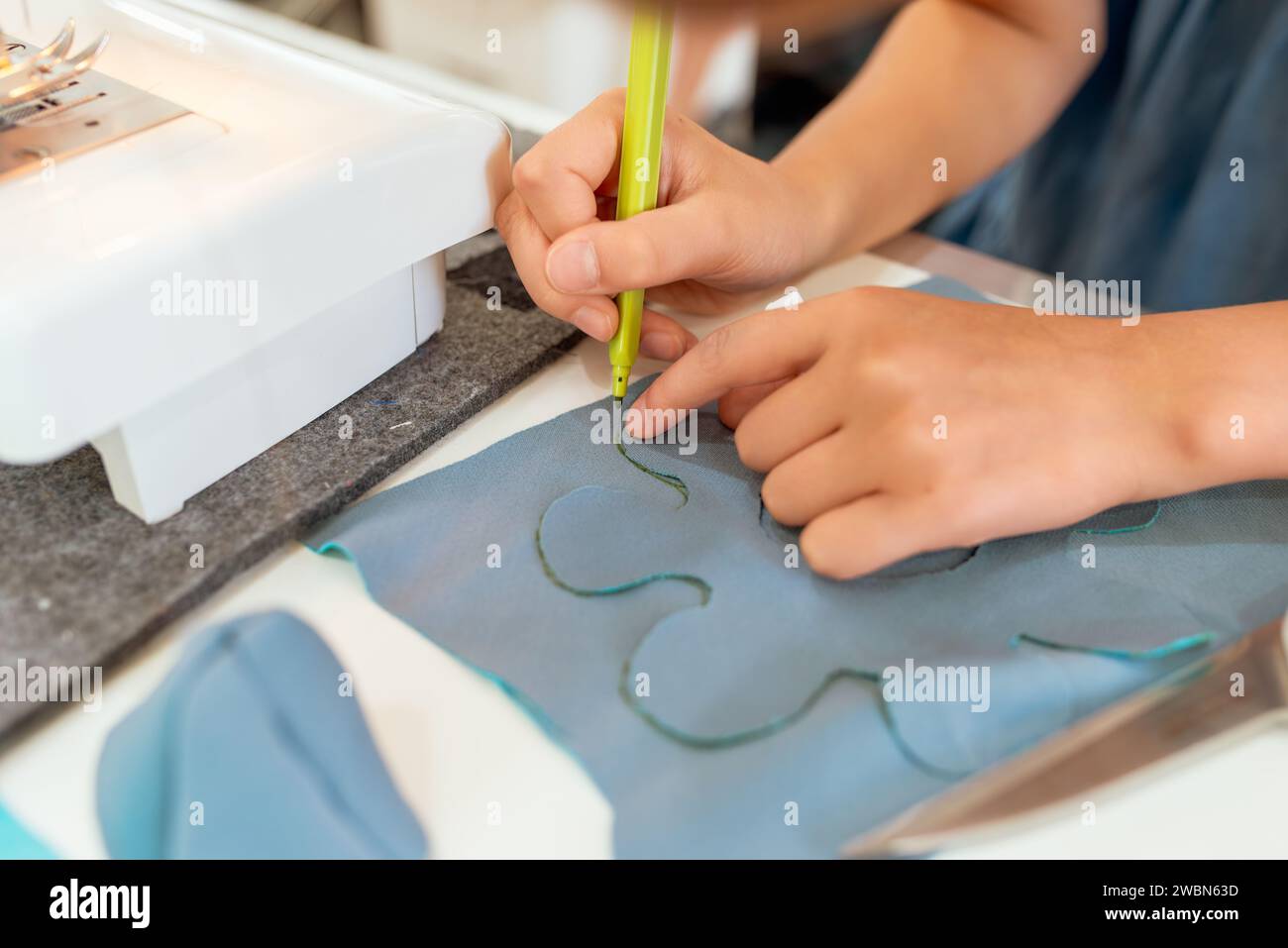 Primo piano di mani femminili che lavorano su una macchina da cucire. È seduta al tavolo e disegna con una matita verde. Laboratori creativi di sartoria per bambini Foto Stock