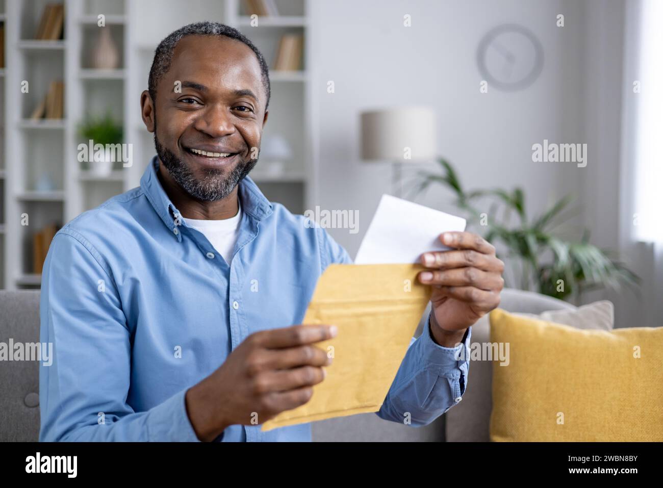 Ritratto ravvicinato di un uomo afro-americano sorridente seduto a casa e con una busta aperta con una lettera in mano, che guarda e sorride la fotocamera. Foto Stock