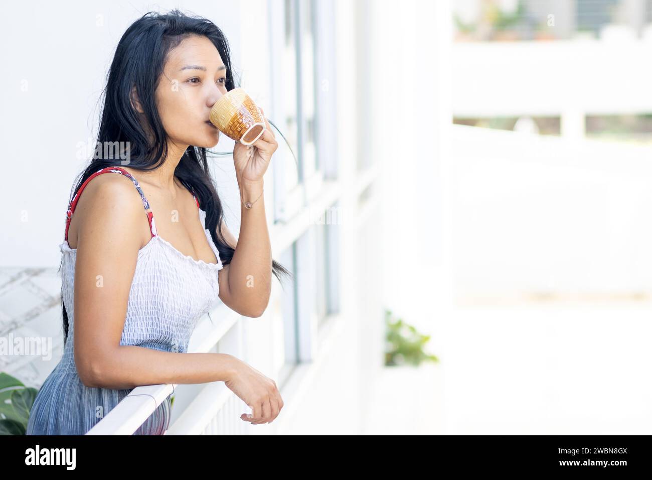 Una donna in abito estivo beve da un tè sul balcone di un condominio in una zona residenziale Foto Stock