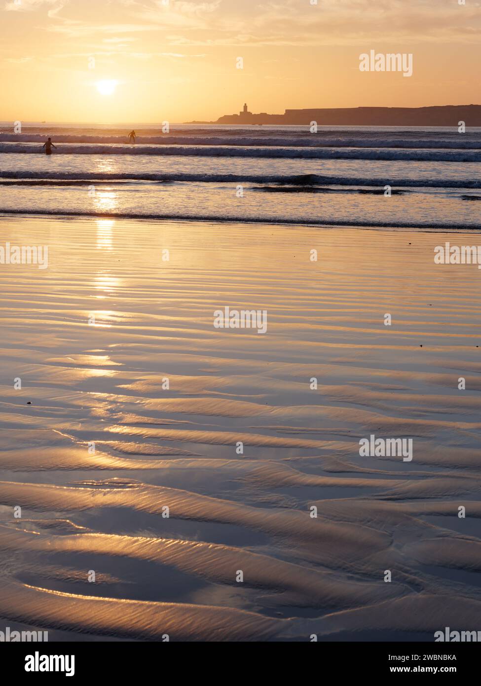 Surfista in mare al tramonto con un'isola alle spalle mentre il sole si riflette nell'acqua e nella sabbia bagnata sulla spiaggia di Essaouira, Marocco, 11 gennaio 24 Foto Stock