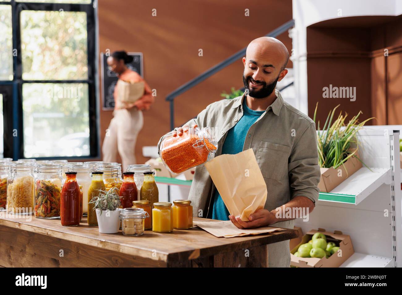 Uomo sorridente versando lenticchie arancioni in sacchetto, acquistando prodotti sani da vasetti di vetro riutilizzabili con prodotti sfusi biologici. Cliente che acquista prodotti biologici naturali presso il negozio locale eco zero rifiuti. Foto Stock