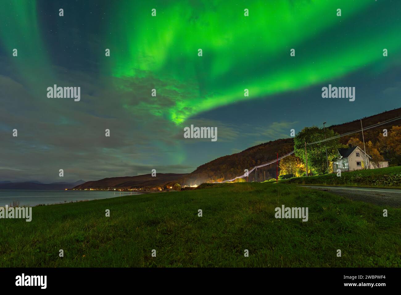 Splendida aurora boreale verde sopra un fiordo e una casa sull'isola di Kvaloya vicino a Tromsø. luci polari danzanti su una montagna, aurora boreale Foto Stock