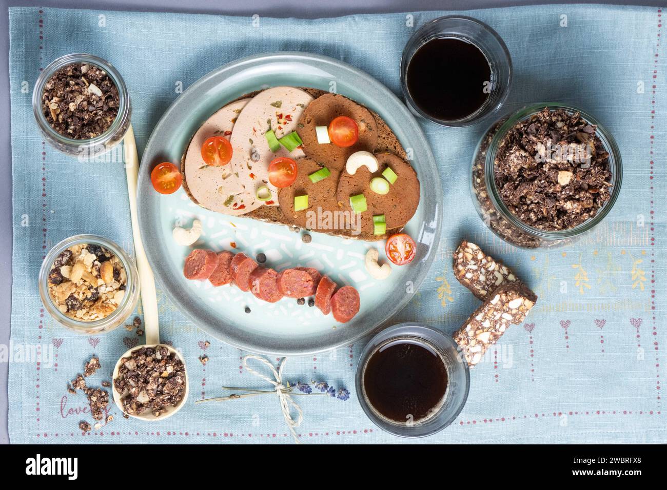 Dresda, Germania. 6 gennaio 2024. Una fetta di pane condita con varie salsicce vegane, una salsiccia all'aglio vegan e una ciotola con un'alternativa di salsiccia al fegato a base di piselli vegani, muesli con yogurt a base vegetale, barretta di datteri di anacardi e caffè su un tavolo. Credito: Sebastian Kahnert/dpa/Alamy Live News Foto Stock