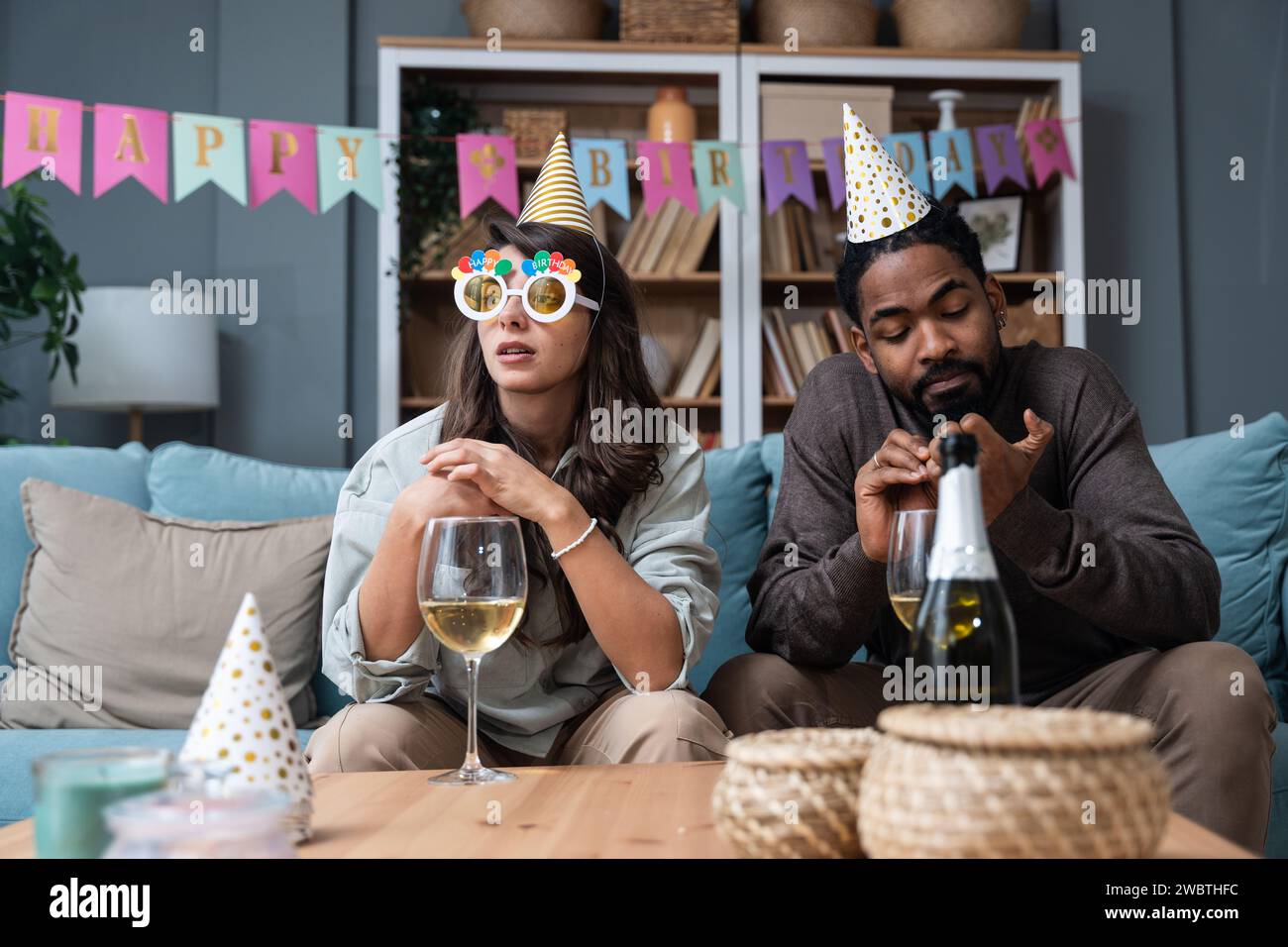 Due giovani seduti su un divano con cappellini di compleanno, triste perché nessuno è venuto a festeggiare il compleanno e la festa nel loro appartamento. Uomo deluso e.. Foto Stock