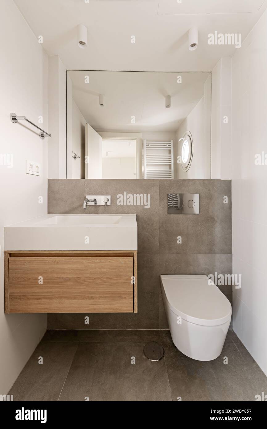 Un piccolo bagno con mobili in quercia, specchio senza telaio, rubinetti di design e WC sospeso Foto Stock