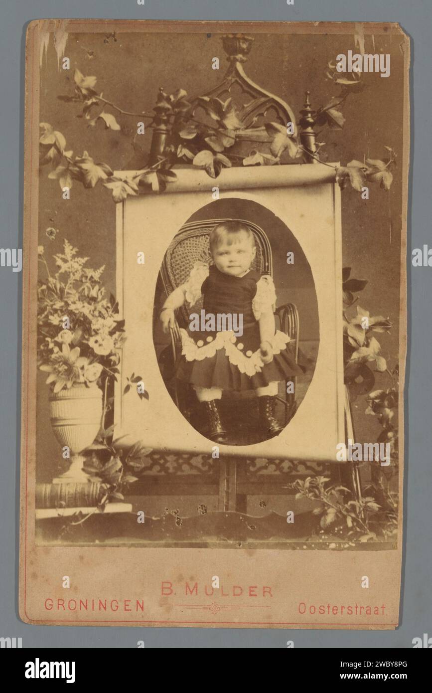 Ritratto di un bambino piccolo su una sedia, Berend Mulder, c. 1880 - c. 1900 Fotografia. Foto cabinet è posizionata su standard con fiori, probabilmente in memoria del bambino. supporto fotografico. stampa di albumi di cartone bambino (a casa) Foto Stock