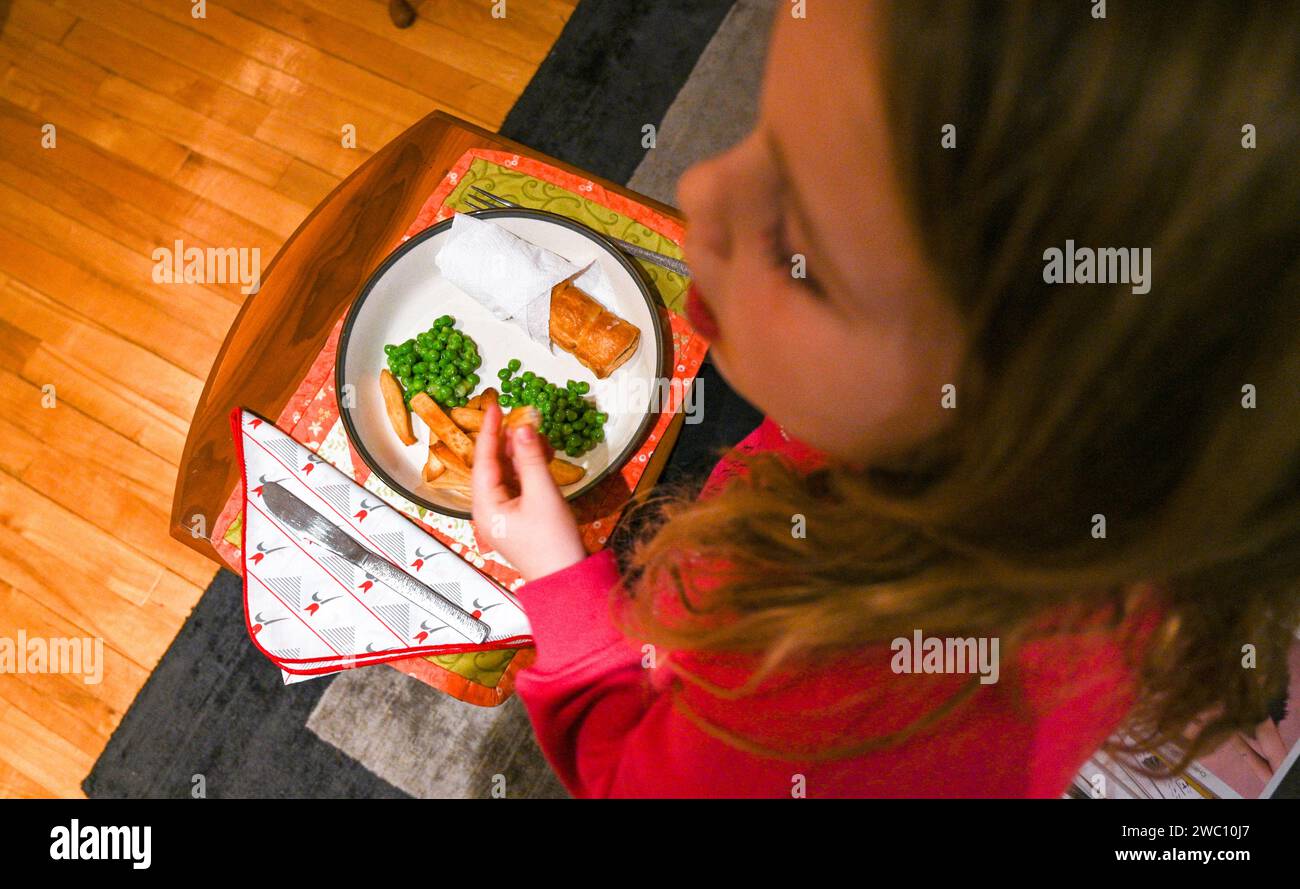Bambina di 7 anni che mangia salsiccia, patatine fritte e piselli Foto Stock