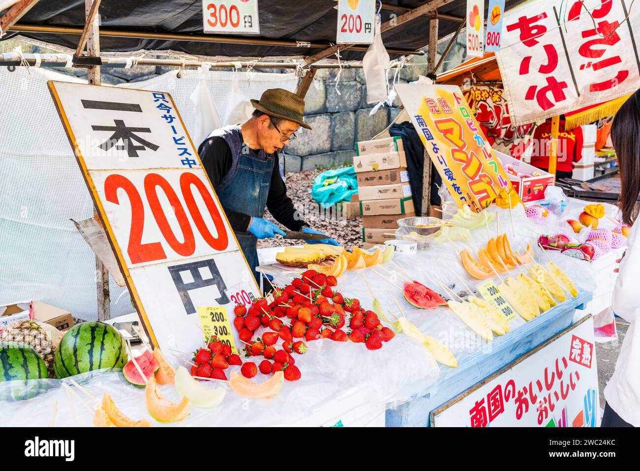 Uomo che taglia pozioni di ananas in una bancarella di frutta con fragole e fette di melone sul bancone del parco del castello di Osaka in primavera. Foto Stock