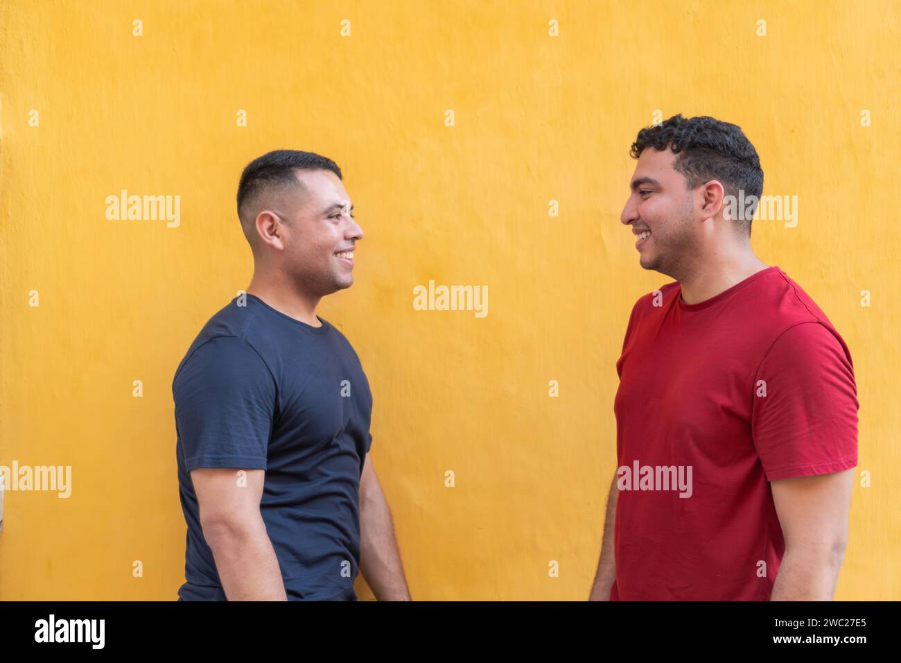 Due uomini in abbigliamento casual, uno in camicia scura e l'altro in rosso, si godono una conversazione leggera con una parete gialla brillante dietro di loro. Foto Stock