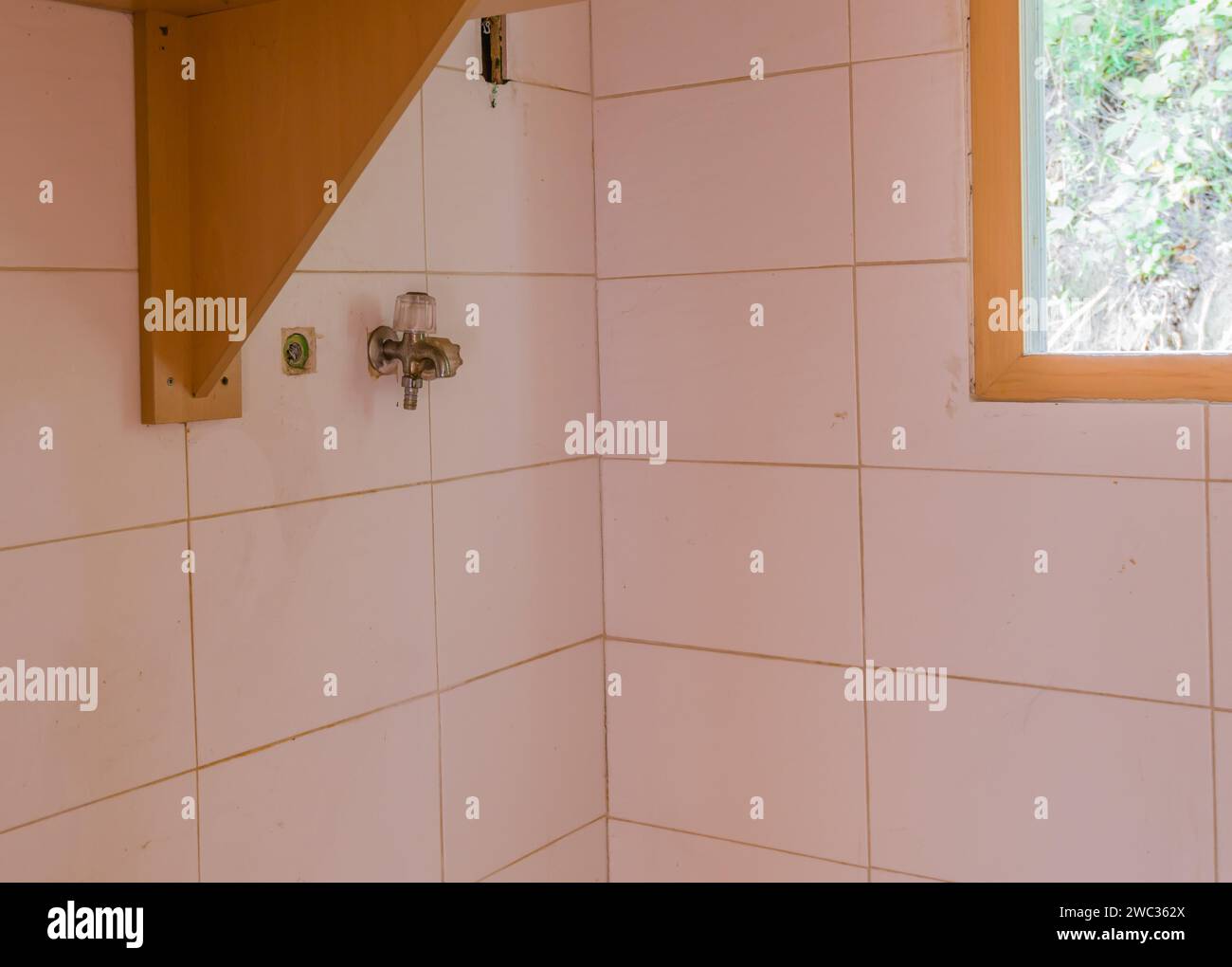 Rubinetto d'acqua su pareti piastrellate bianche accanto a scaffale e finestra in legno nella lavanderia della casa abbandonata Foto Stock