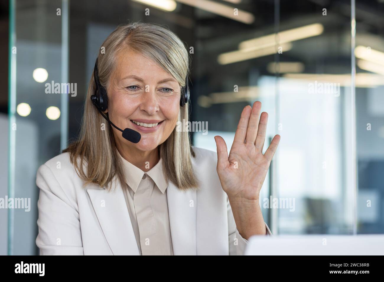 Donna matura professionale con cuffie che salutano in modo amichevole, rappresentando l'assistenza clienti e l'interazione positiva sul luogo di lavoro. Foto Stock
