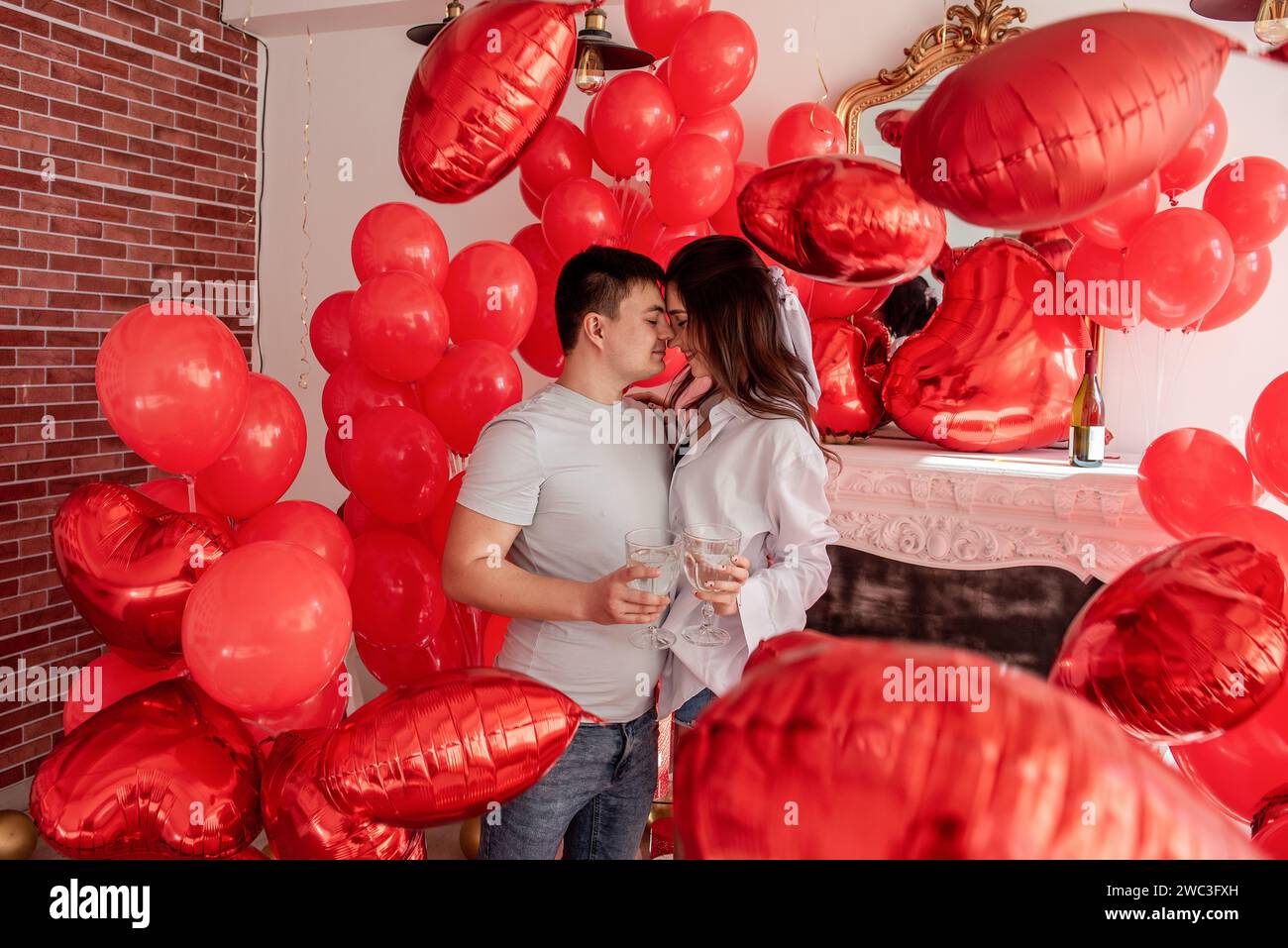 Un momento di gioia tra giovani coppie che festeggiano con un brindisi di San Valentino vicino a palloncini rossi, caminetto bianco. Donna che ride, tiene il bicchiere, sta per C. Foto Stock