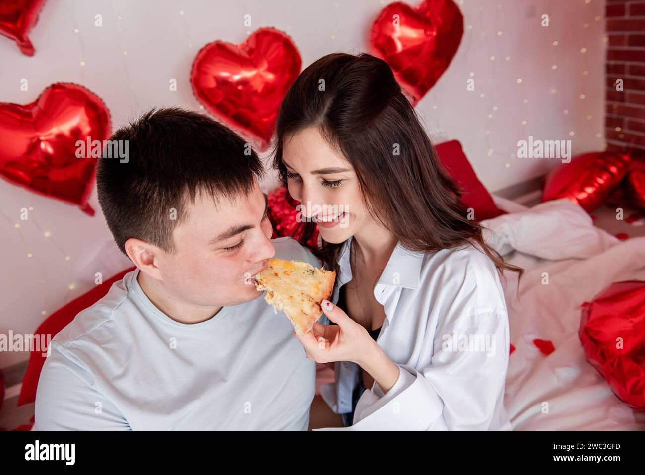Primo piano di allegri coppie innamorate che si nutrono a vicenda della pizza sedendosi su un letto bianco tra palloncini a cuore rosso il giorno di San Valentino. Appuntamento informale con Foto Stock