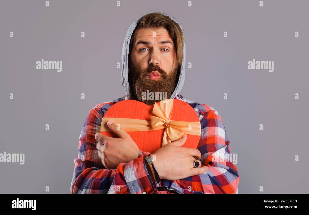 Buon San Valentino. Uomo barbuto con scatola regalo rossa a forma di cuore che manda baci d'aria. Shopping di San Valentino. Bel ragazzo a forma di cuore Foto Stock