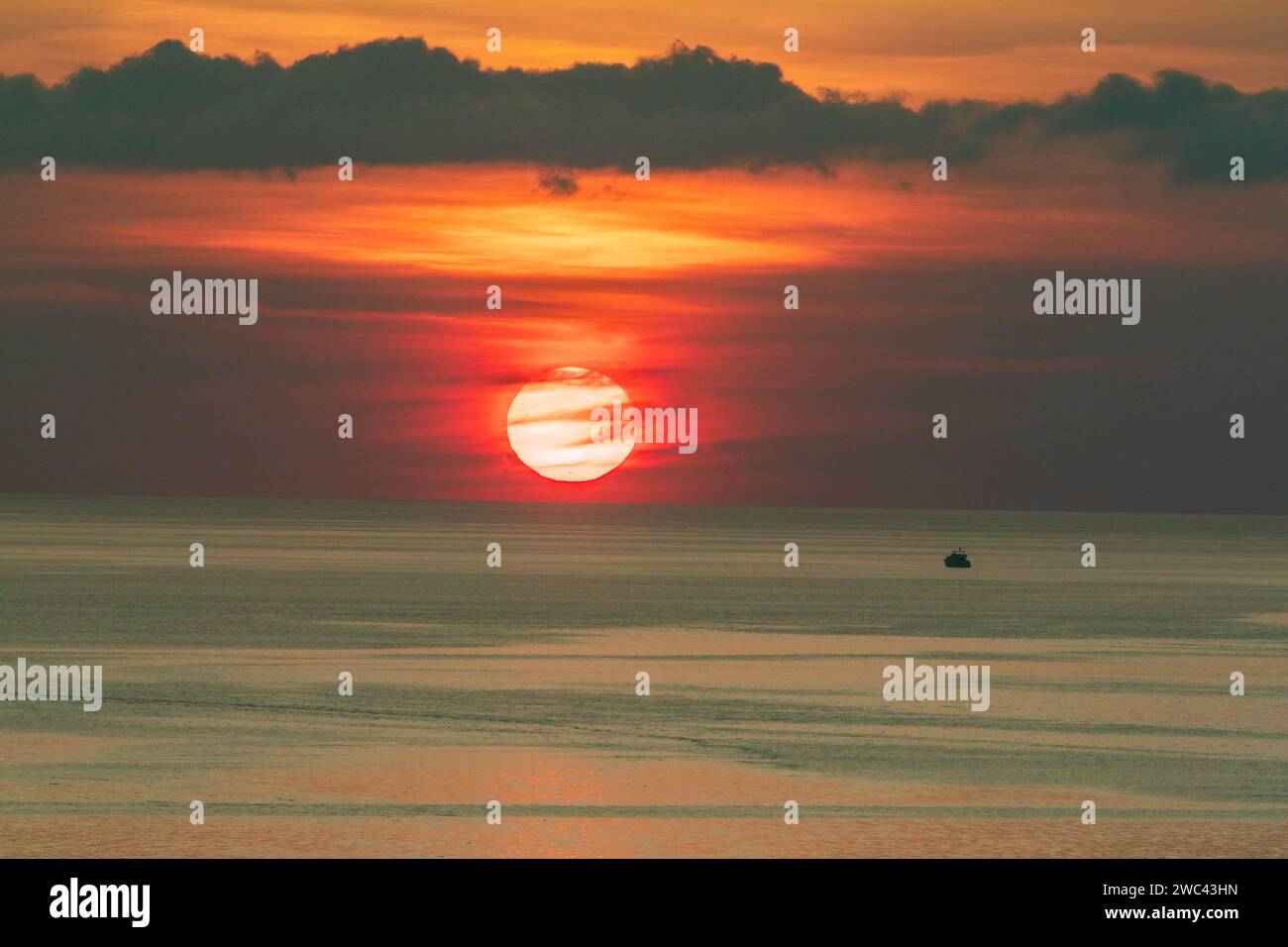 Grande sole rosso e arancione che tramonta sulla linea dell'orizzonte oceanico e piccola barca da pesca Foto Stock