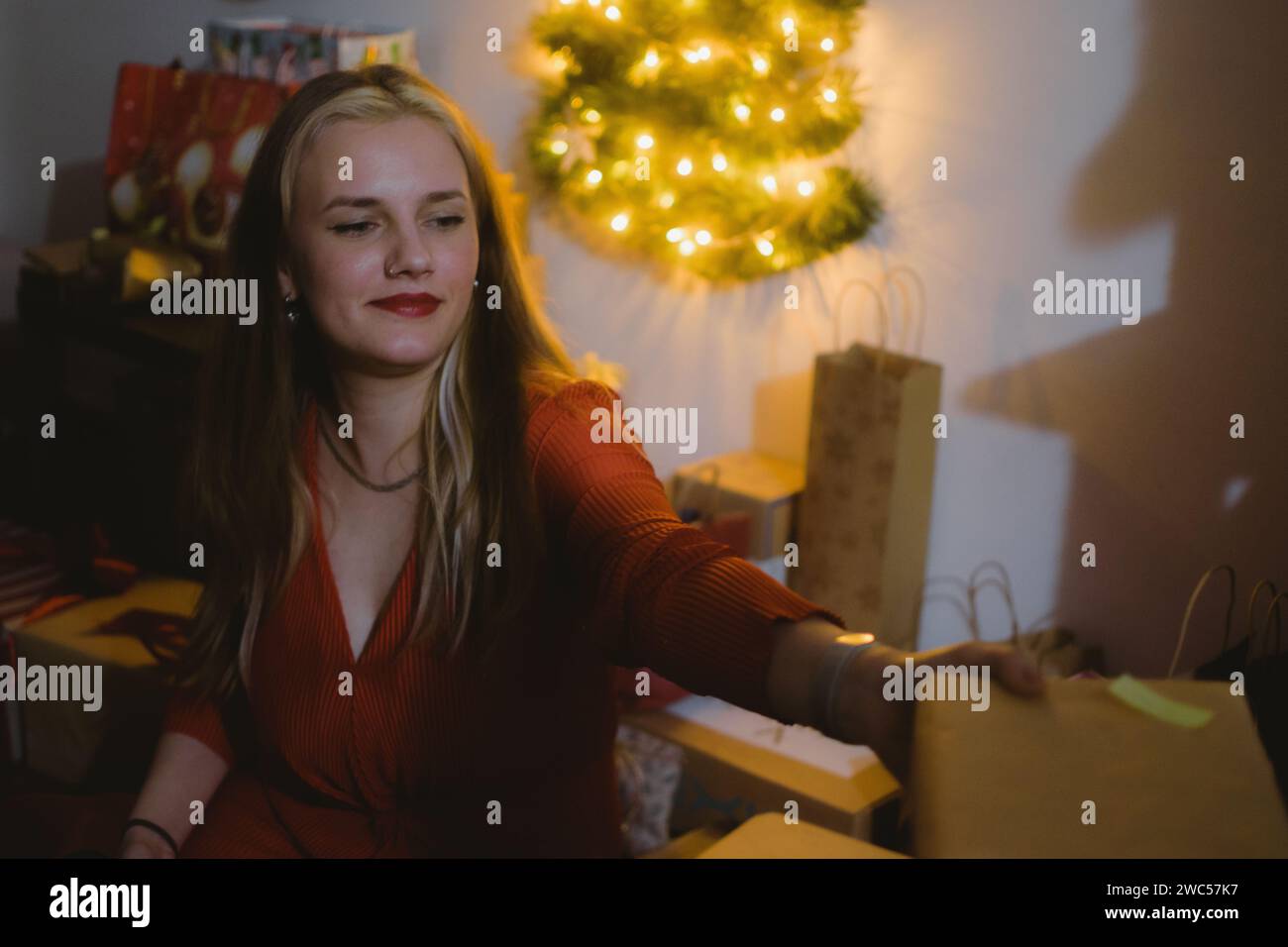 Una donna gioiosa che scarta con ansia una confezione regalo accanto a un albero di Natale splendidamente decorato illuminato da luci vibranti Foto Stock