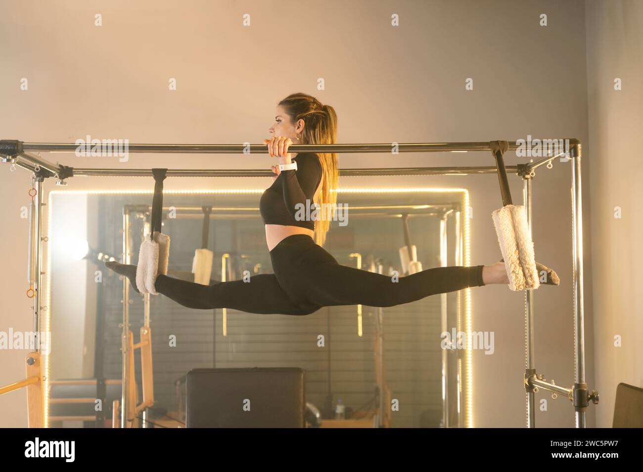Donna esegue un esercizio di flessibilità avanzato su un riformatore Pilates, sospendendosi con le gambe separate Foto Stock