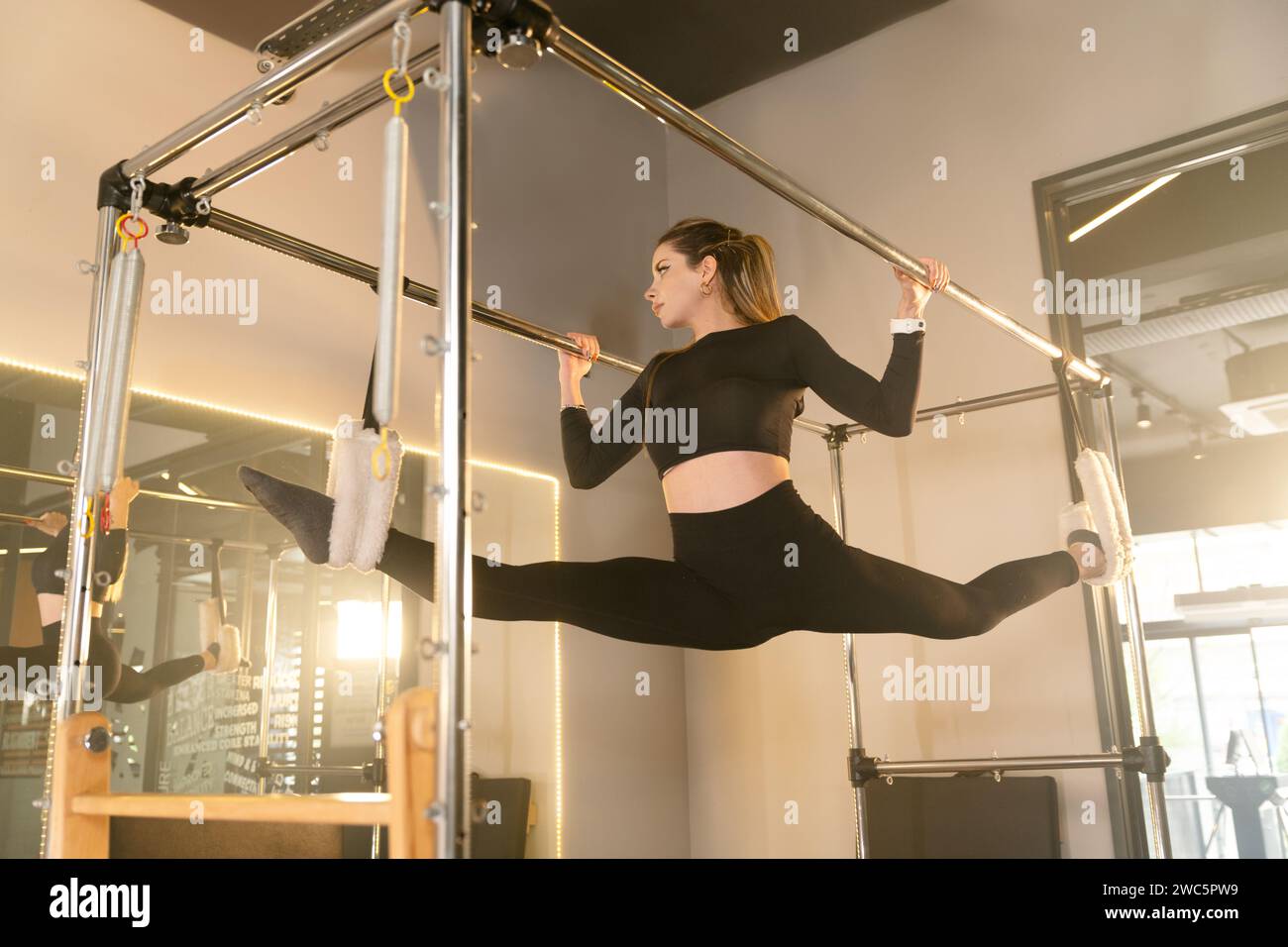 Donna esegue un esercizio di flessibilità avanzato su un riformatore Pilates, sospendendosi con le gambe separate Foto Stock