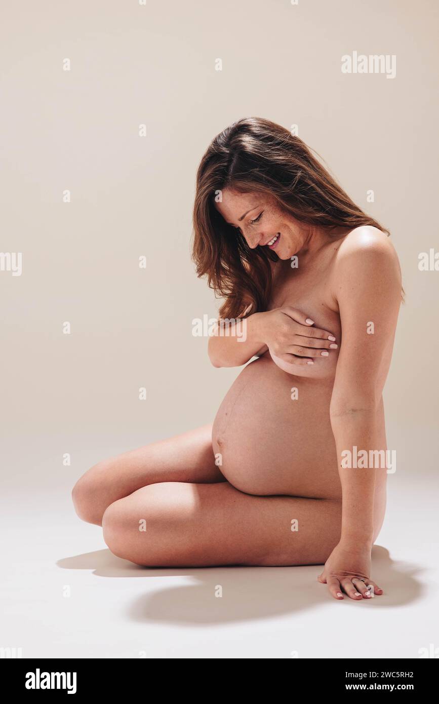 Felice donna incinta nuda al terzo trimestre siede sul piano dello studio, sorridendo al suo batto. Aspetta mamma che festeggi la sua gravidanza e mothe Foto Stock