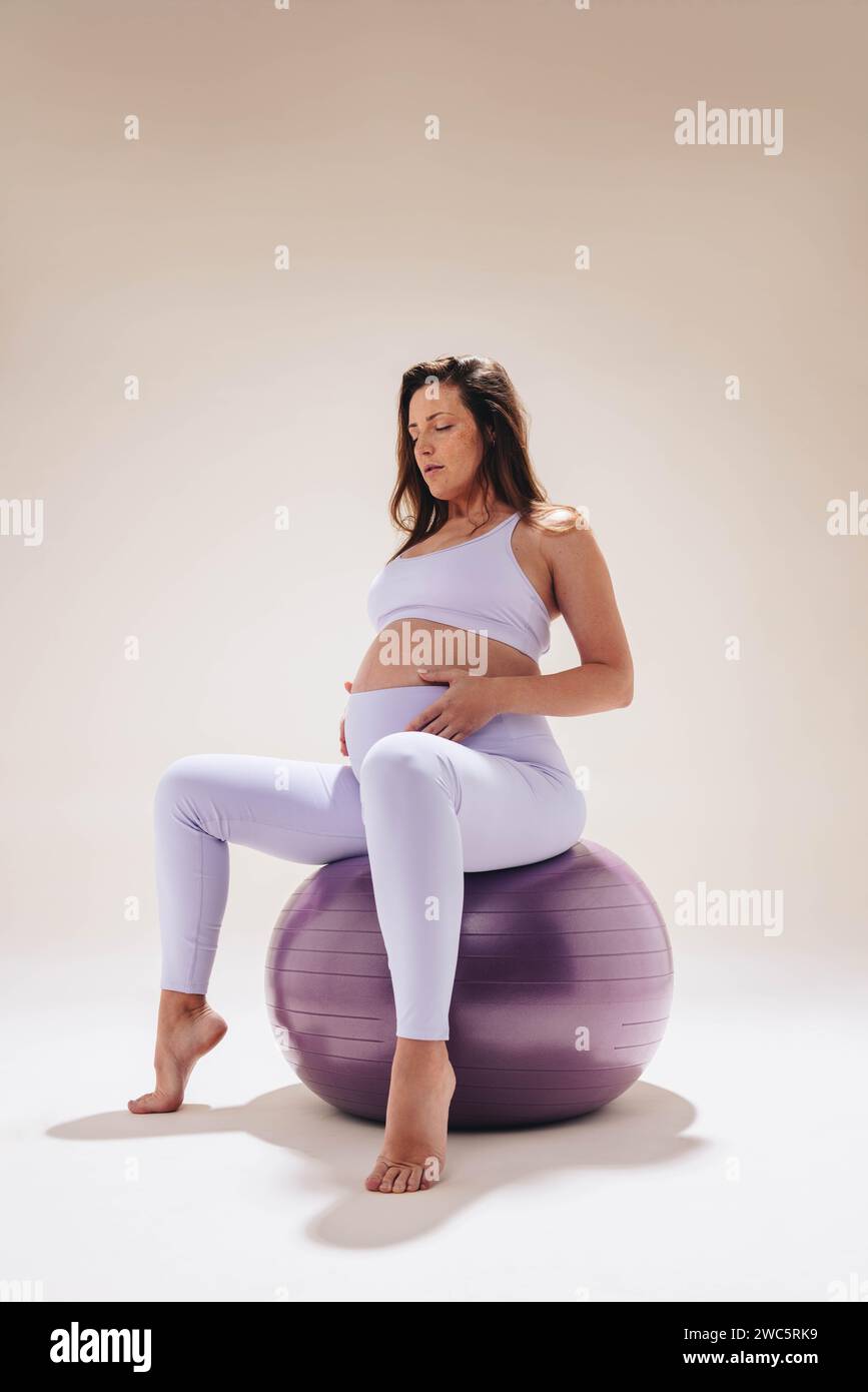 donna incinta nel suo terzo trimestre pratica yoga prenatale in uno studio. Utilizza una palla da ginnastica per la stabilità e la flessibilità, concentrandosi sull'auto-c Foto Stock