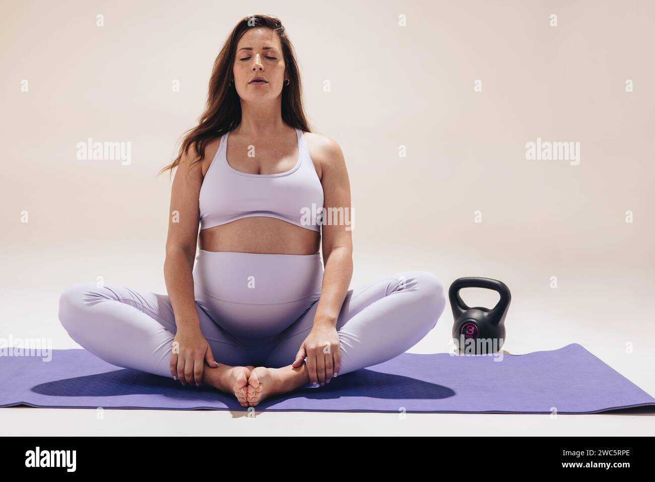 Una donna serena e incinta pratica yoga in uno studio. Si siede su un tappetino per esercizi, con il suo piccolo batto visibile, in posa a farfalla. Concentrata sul suo respiro Foto Stock